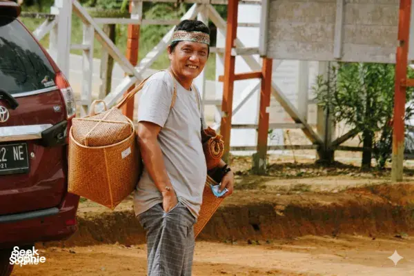 Riyan Momod, local host, stands outdoors in Pangkalan Bun holding a woven basket, with a parked red SUV beside him