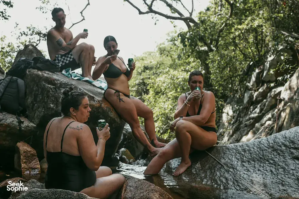 Travellers relaxing on rocks by a jungle stream, holding drinks during a Hoi An secret waterfall motorbike adventure