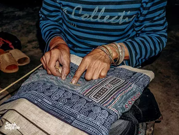 Hands tracing an intricate design onto patterned fabric in a textile craft workshop in Vietnam, striped sleeves visible