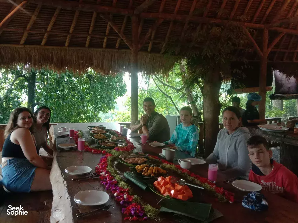 Guests enjoying a communal dinner on a long green table runner with flowers and candles under a thatched roof at Omunity Bali, Bali