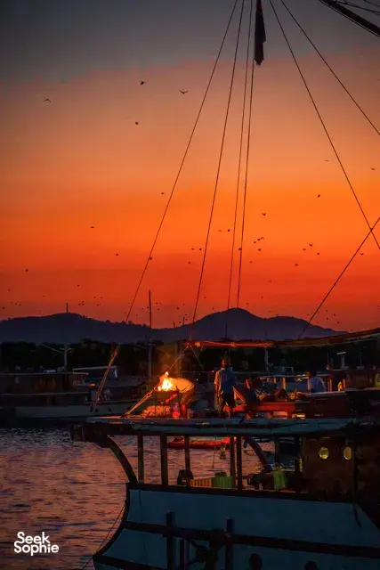 A boat deck at dusk with a small fire and fruit bats circling overhead, sails and ropes frame the sky, Komodo Island