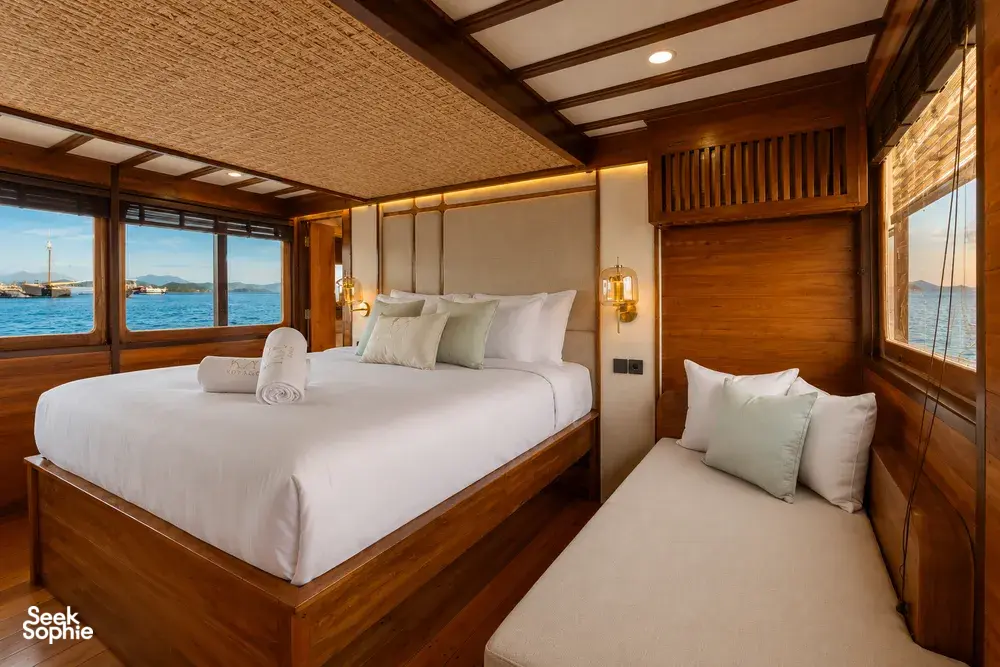Travass Life phinisi cabin with a double bed, lounge seating and sea-window views for a Komodo shared trip, Labuan Bajo