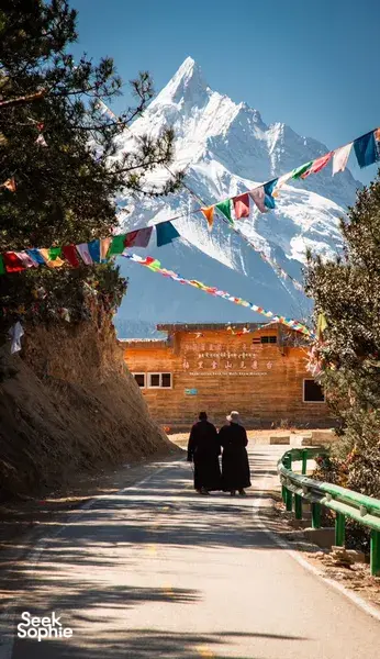 Travellers walking a sunlit road lined with prayer flags toward a snow-capped peak on the Shangri-La Loop near Shangri-La