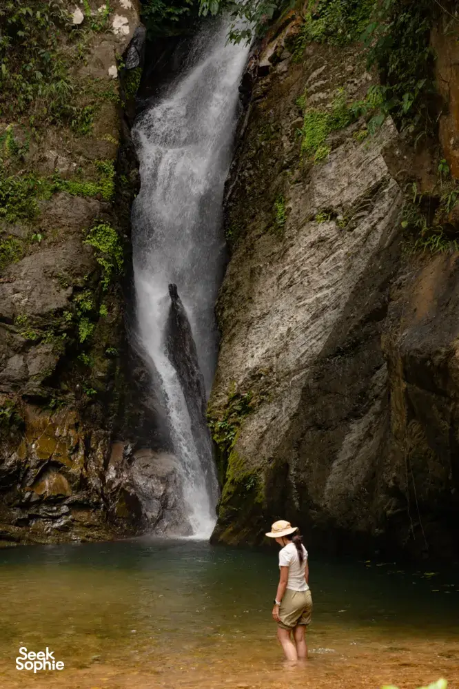 Traveller in a cap standing near a tall waterfall pouring into a rocky pool during a Borneo Overland wild Sabah road trip