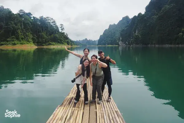 Friends posing on a bamboo raft in Khao Sok National Park, cruising past towering limestone cliffs on a calm lake