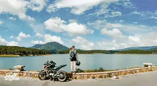 Traveller standing with parked motorbikes by a lake railing, with hills and blue sky in the background on an Easy Rider trip