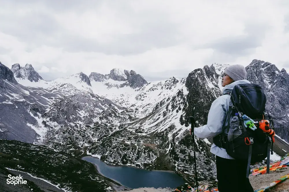 Trekker in a beanie with a large backpack gazing over snow-dusted mountain valleys and a turquoise lake in Shangri-La