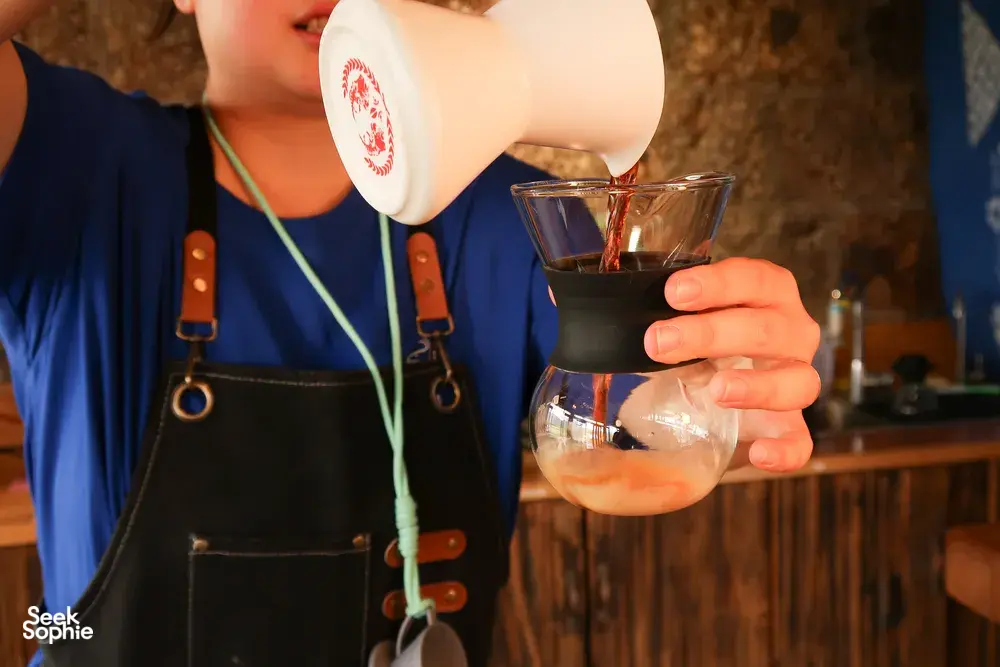 A pour-over moment in a Lijiang coffee workshop, as a taster pours brewed coffee into a glass cup for tasting