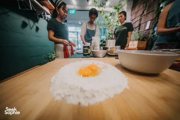 Flour ring and raw egg on a wooden pasta table as others prepare ingredients for workshops in Singapore