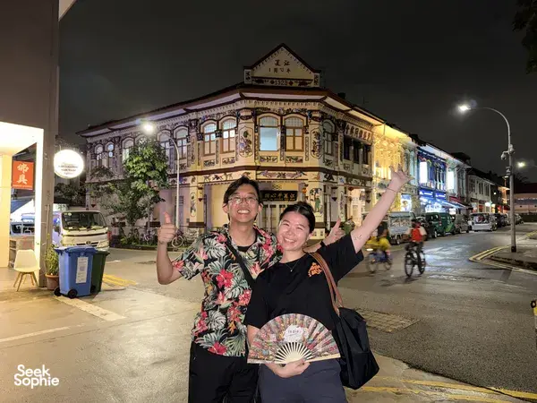 Two people posing on a night street in Geylang, Singapore, holding a folding fan in front of a heritage shophouse