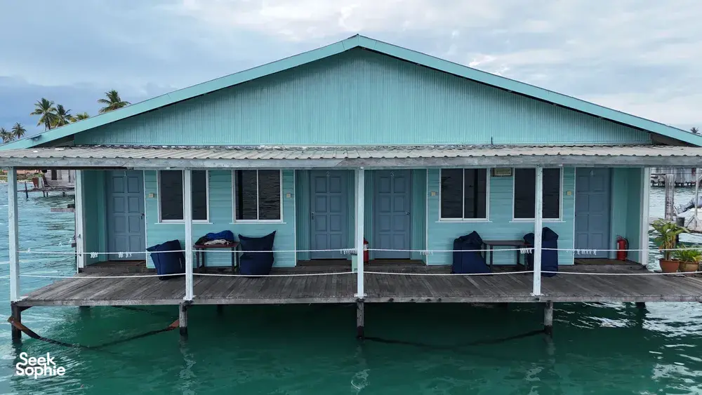 Banglo air kayu berwarna hijau kebiruan di atas tiang di Semporna, dengan tempat duduk di anjung hadapan dan laluan pejalan kaki di atas laut yang tenang