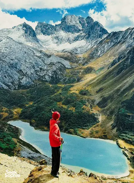 Hiker in a bright red jacket overlooking a turquoise alpine lake and steep valleys beneath rugged mountains in Shangri-La