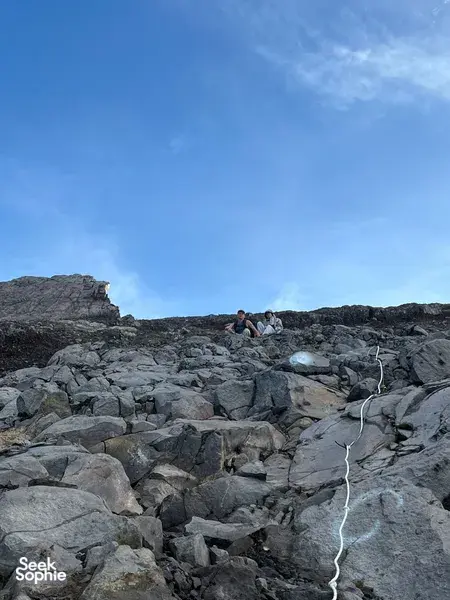 Hikers crouch on Mount Agung’s rocky summit ridge under a bright blue dawn sky during a private sunrise trek with local guides
