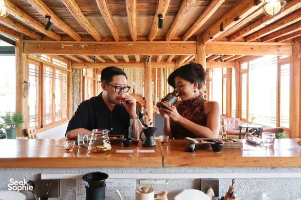 Lijiang coffee workshop with a wooden counter and long windows, two people sipping and tasting brewed coffee