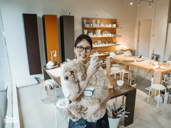 Ina Atelier host smiling and holding a small dog in a bright ceramics studio with shelves of handmade dishes, workshops in Singapore