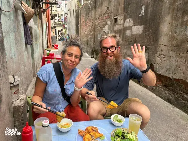 Smiling travellers waving from a street-feast table in a narrow Hanoi alley with bowls of food and drinks
