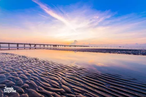 Sandy beach at dusk with footprints leading to a long pier and calm water, with a sky of soft pastel sunset