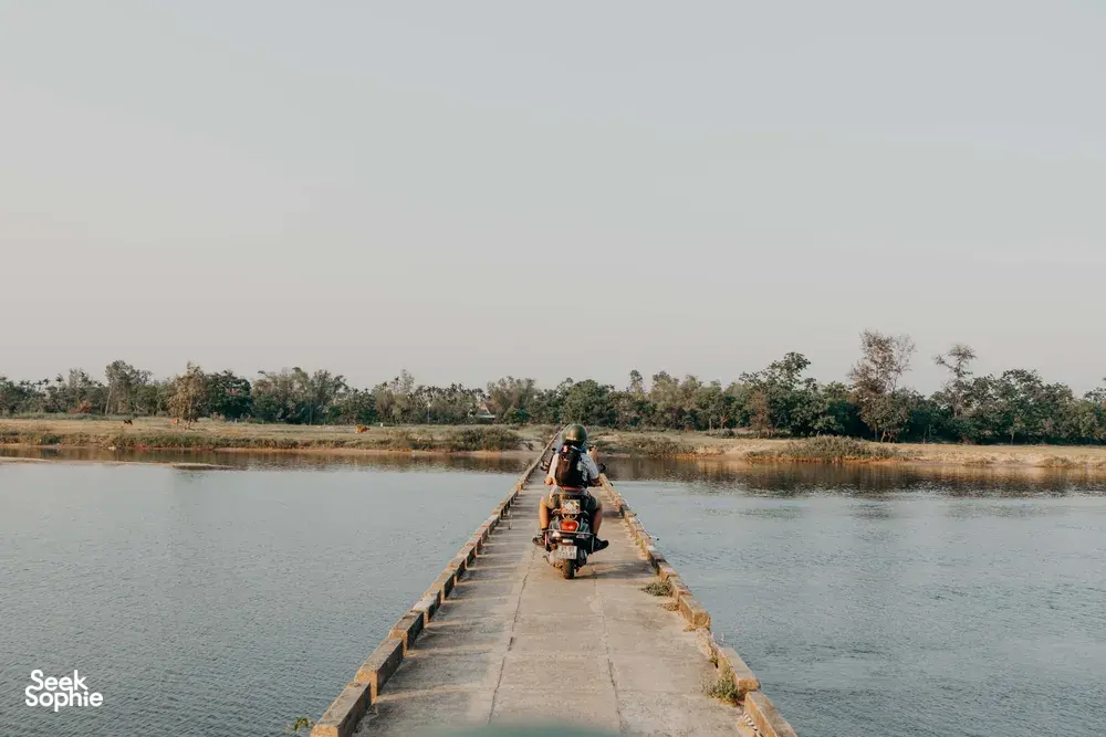 Sebuah skuter melintasi jembatan panjang di tepi sungai, salah satu sorotan rute jalan kecil pilihan Hoi An Backroad Tours, dekat sungai yang tenang di Hoi An.