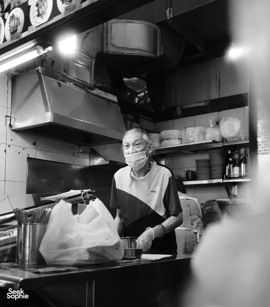Black-and-white view of an elderly hawker cook preparing food in a Singapore kitchen stall beneath vent hoods and shelves