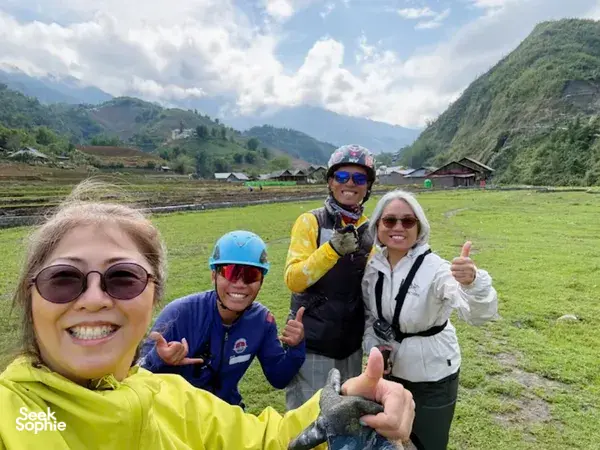 Group of paragliding passengers in helmets smiling and giving thumbs up on a grassy launch field in the hills, Sapa