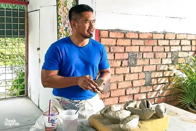 Ceramic artist in Ilham Ceramic Studio, Langkawi, shaping pottery at a table outdoors beside a brick wall