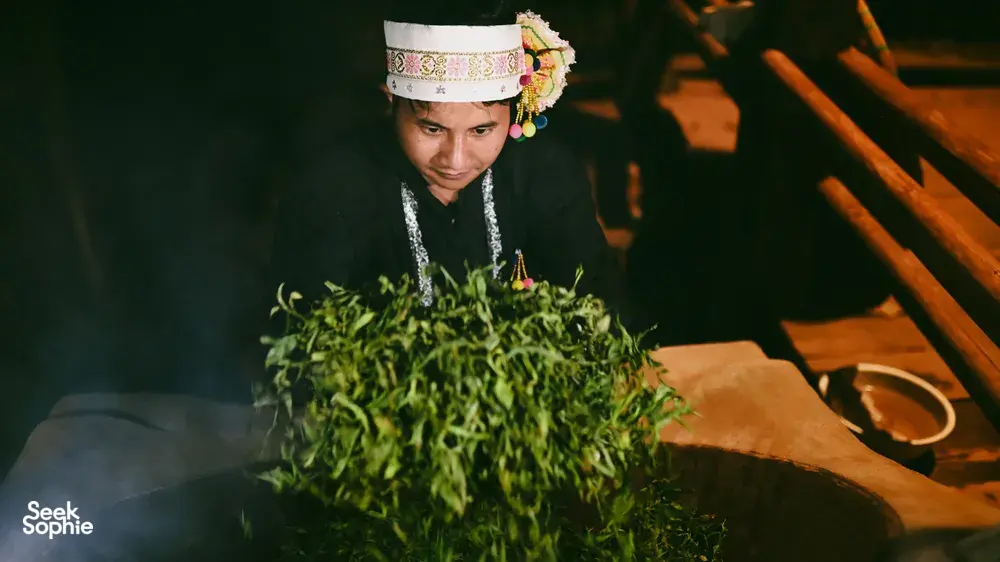 Seorang perempuan setempat dengan hiasan kepala bordir memegang seikat herba hijau segar di ambang pintu desa di Xishuangbanna