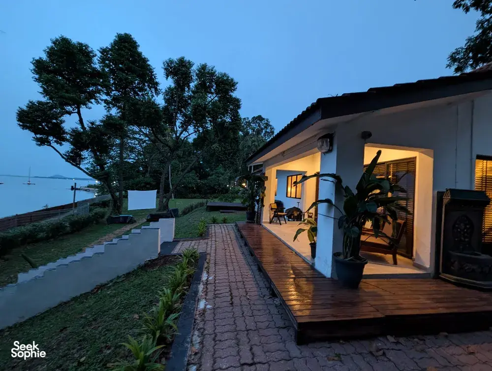 Dusk view of a wooden deck with chairs beside large windows, garden paths leading to the sea at a Singapore 2D1N farmstay