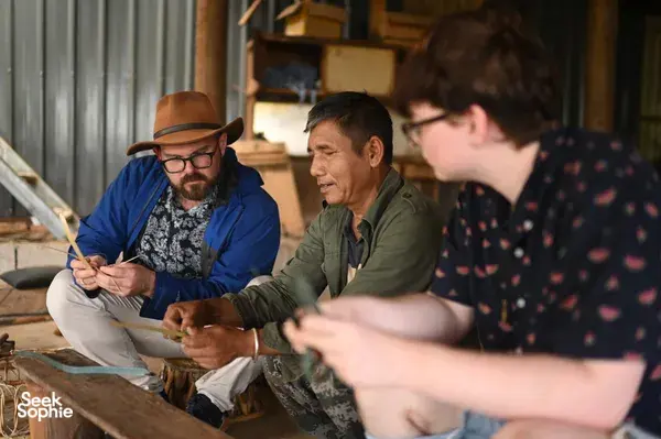 Local guide from Tea Origins helping travellers practise traditional tea-making by a wooden table in a village workshop, Xishuangbanna