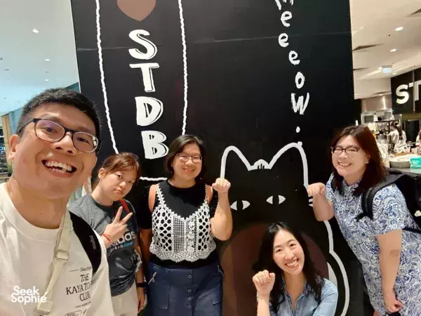 Smiling team posing in a Singapore indoor game room beside a cat-themed board for “Secrets of Wealth” — team building Singapore