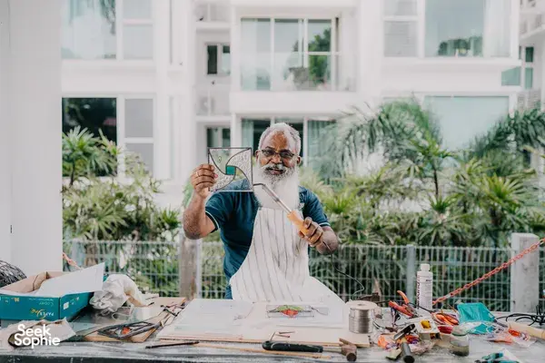 Elderly glass artist shaping a curved stained-glass panel at a table in a bright garden, Singapore—workshops in Singapore