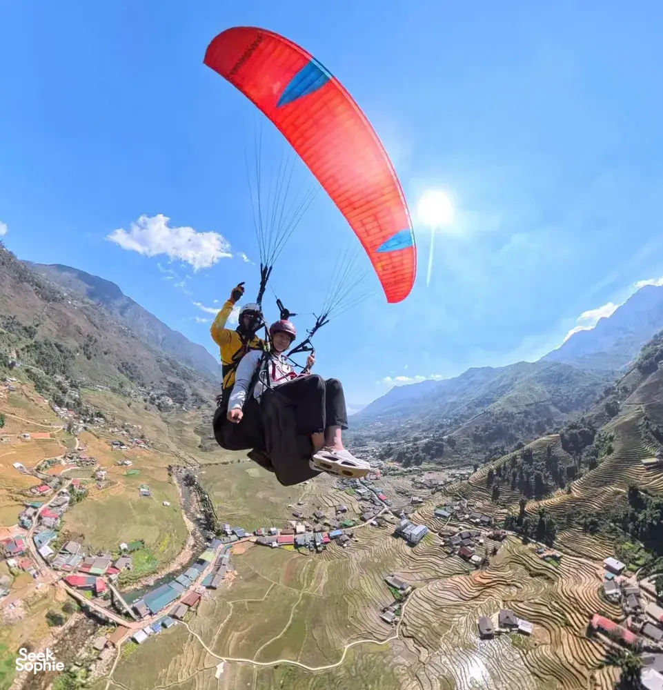Two paragliders floating over patchwork terraces and a winding river, red-orange canopy catching the wind above the mountains in Sapa