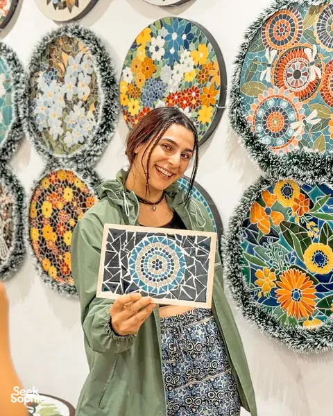Smiling traveller holding a finished ceramic mosaic panel at a Bat Trang workshop near Hanoi, with round mosaic artworks behind