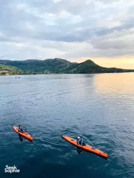 Two orange kayaks with paddlers on calm water at sunset in Labuan Bajo, with green hills fading into the sea at dusk