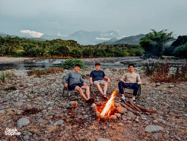 Friends relaxing around a campfire on a rocky riverbank with forested hills and mountains in Sabah near Kota Kinabalu