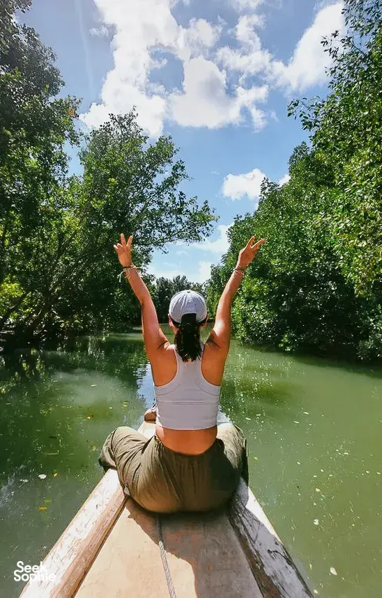 Woman sitting on a boat deck with arms raised above calm green water and thick trees during a mangrove visit in Labuan Bajo