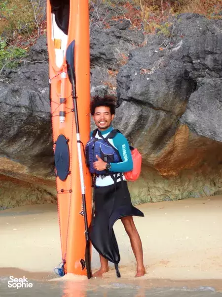 Smiling paddler standing in shallow surf beside an orange kayak at Labuan Bajo, holding a paddle near a rocky shore after kayaking