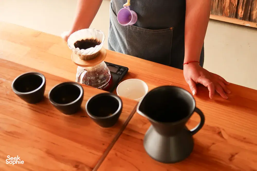 Coffee tasting table in Lijiang with black sample cups, a glass grinder jar, and a pour as visitors try Yunnan brews