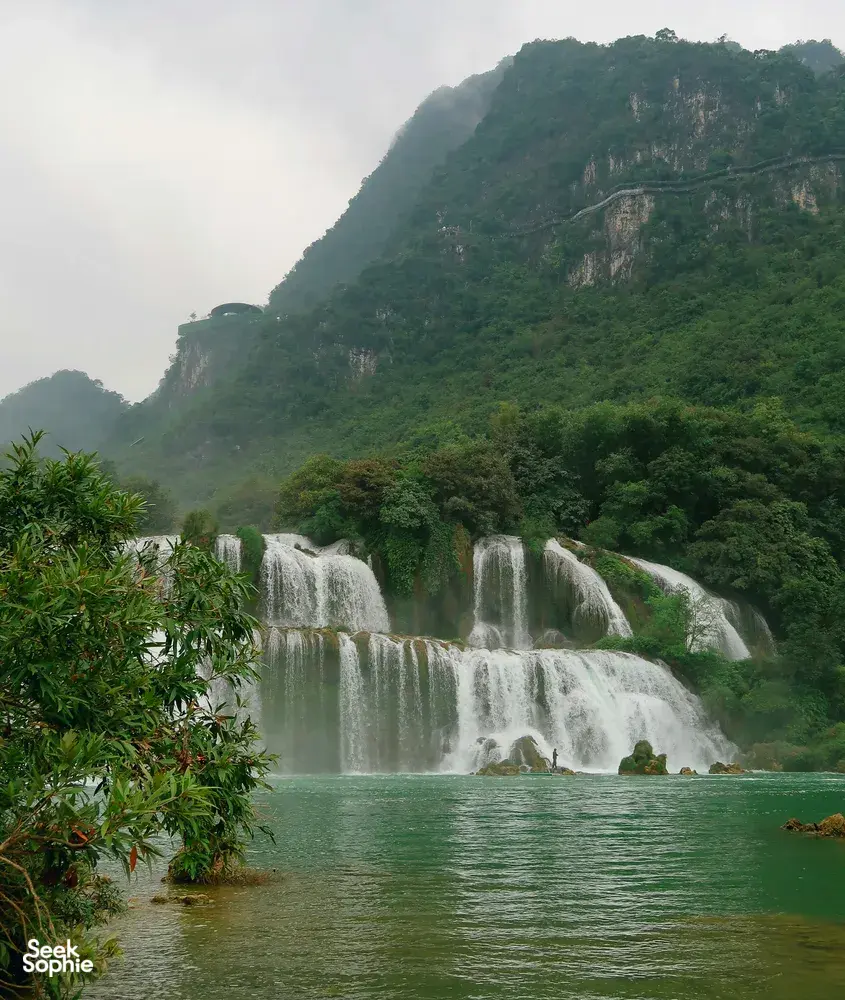Tiered waterfalls flowing into a green river with mist, backed by steep forested mountains under an overcast sky
