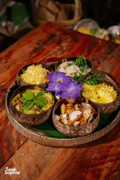 Coconut-shell Thai dishes served on a wooden table with purple flowers and herbs, part of a local farm meal
