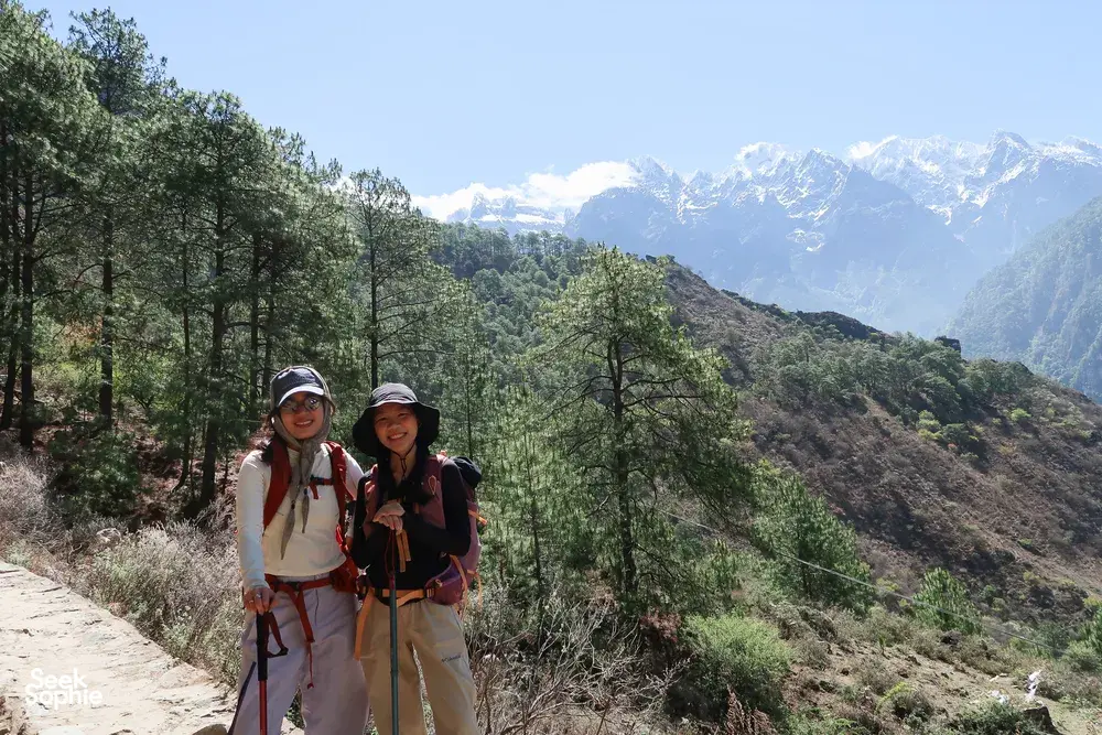 Two hikers on a dusty mountain footpath with pine trees and distant peaks during the Tiger Leaping Gorge high trail, Lijiang