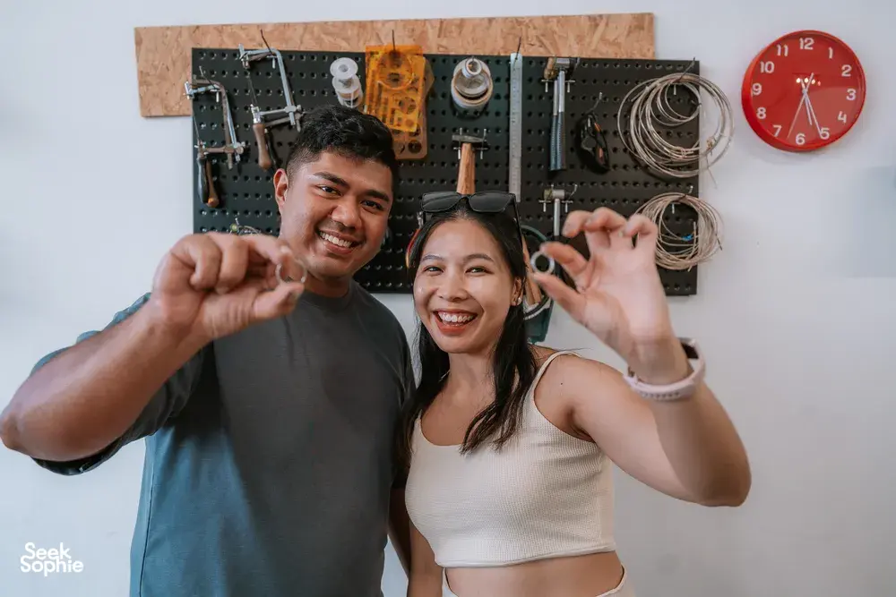 Smiling couple holding handmade rings in front of a wall packed with jewellery-making tools at Fat Anvil Studios, Singapore