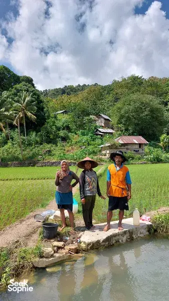 Persinggahan di kampung tepi sungai dalam lawatan sehari di Labuan Bajo: tiga orang bergambar di tepi terusan, dengan sawah berteres yang menghijau dan rumah bertiang