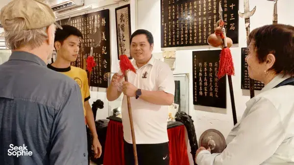 A guide in a white polo explains a lion dance instrument while guests stand around red drapes and calligraphy in Ho Chi Minh City