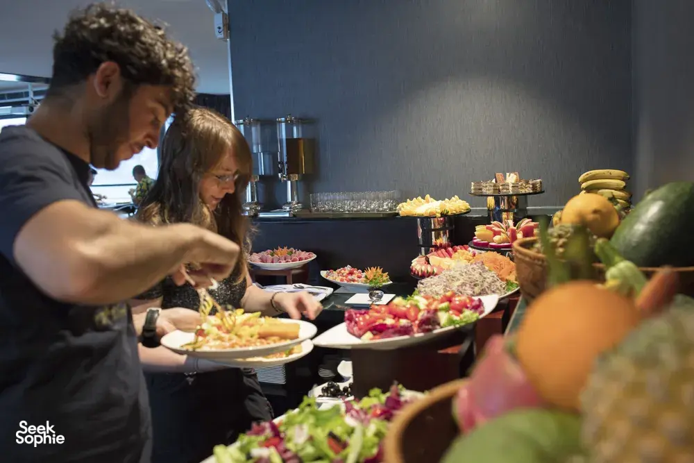 Guests enjoying a buffet on a party cruise, with fruit and appetisers arranged on serving stations in Halong Bay