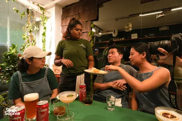 Guests chatting and tasting pizza at Secret Pizza Club in Singapore, while a host gestures near a wood-fired oven in a cosy indoor space