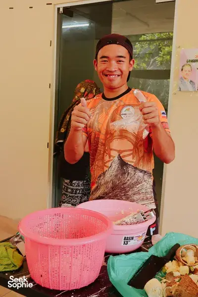 Smiling young man holding a wrapped, home-cooked dish in a village doorway beside a pink basket, Kota Kinabalu