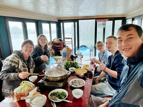 Friends smiling over hotpot dining on a Cat Ba boat, with large windows showing calm bay water and limestone hills