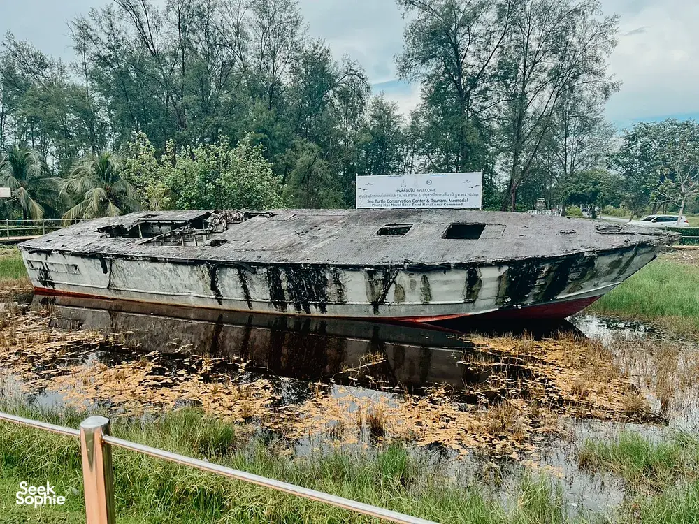 Old, damaged boat resting in shallow muddy water and grass, with trees and a sign in a quiet riverside area