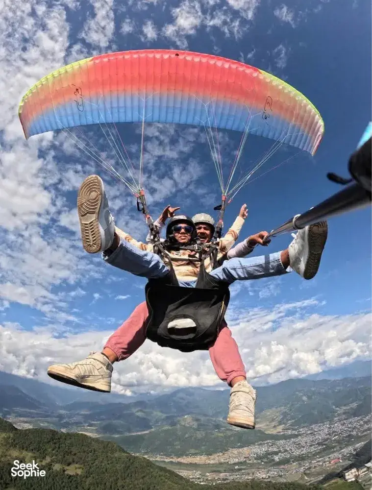 Paraglider rider soaring under a rainbow canopy with legs extended and friends in harnesses above a vast mountain valley, Sapa