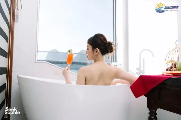 Sunshine Golden Cruises guest relaxing in an onboard bath tub on a Cat Ba cruise, holding an orange drink by the window